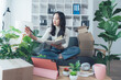 © Kritdanai - A Moment of Serenity: In a sunlit home office, a young woman finds balance amidst the lush greenery. Dressed casually, she appears focused, perhaps sketching or jotting ideas in her notebook.