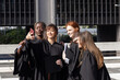 © wavebreak3 - Diverse female graduates posing for selfie on plaza wearing gowns, holding phone, diplomas, cap