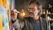 © swillklitch - Man in early 40s with thin frame glasses writing on colorful sticky note attached to corkboard promoting neurodivergent creative brainstorming process