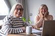 © luciano - Two smiling senior women with white hair and glasses sitting at a wooden table at home, sharing laptop together. They appear relaxed and happy while browsing online, with papers and a coffee mug
