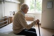 © raquel - Lonely elderly man sitting on hospital bed with walking cane in nursing home room reflecting aging and healthcare support