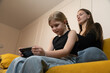 © Volha - Young girl focusing on playing a mobile game on her smartphone, sitting on a yellow couch with her older sister observing her at home, enjoying leisure time together braiding her hair