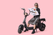 © Pixel-Shot - Beautiful young fashionable woman in trendy outfit with electric scooter and newspaper on pink background