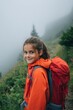 © rabbit75_fot - Explore adventure outdoor in National Park. A young girl in a bright orange jacket standing on a grassy hillside, carrying a backpack. The background is slightly blurred.