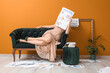 © Pixel-Shot - Young woman in high heels with many newspapers and phone sitting on sofa near orange wall