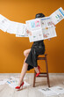 © Pixel-Shot - Young woman in high heels with many newspapers sitting on chair near orange wall