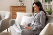 © Pixel-Shot - Young woman in grey pajamas with coffee and laptop working on sofa at home