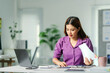 © Crystal - Asian businesswoman working diligently at desk, holding documents, and reviewing information on her laptop in a modern office setup