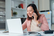 © amnaj - Happy Asian businesswoman making a call, smiling while managing work and communication in a modern office environment