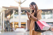 © Art_Photo - Happy asian woman shopper holding paper bags and use smartphone outdoors, Beautiful girl checking online sale promotion or social media on mobile phone in city park, Consumerism and lifestyle concept