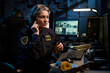 © pressmaster - Middle aged Caucasian woman police officer sitting at desk wearing headset holding coffee cup, working at computer with surveillance monitors and paperwork in dimly lit office