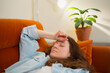 © wifesun - Young woman reclining on an orange sofa with eyes closed and hand to forehead, showing fatigue and discomfort while resting at home, quiet and vulnerable moment of illness