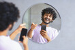 © Prostock-studio - A man stands in a modern room and looks at his reflection in a round mirror. He smiles and points while holding a smartphone. The setting suggests a casual moment of enjoyment and interaction.