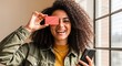 © ArtFocus Studio - Happy young woman with curly hair holding a blank pink credit card over one eye while using her smartphone indoors.