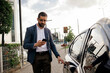 © kleberpicui - Urban professional using smartphone to request or access ride hailing transportation beside a parked car in a modern city environment, representing app mobility and daily commuting.