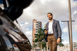 © kleberpicui - Urban professional using smartphone to request or access ride hailing transportation beside a parked car in a modern city environment, representing app mobility and daily commuting.