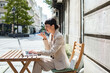 © Marko Geber - Businesswoman using voice assistant while working at outdoor cafe