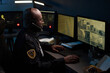 © pressmaster - Black man wearing headset working as security officer monitoring multiple surveillance screens in control room at night, focusing on urban building footage and maps