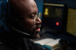 © pressmaster - Black man wearing headset working at computer station, speaking and focusing on screen, glasses reflecting monitor light, blurred background with monitors and notebook visible