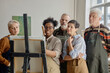 © Seventyfour - Group of middle aged and senior multiethnic men and women observing young Black woman demonstrating painting technique on canvas during educational art class indoors