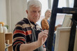 © Seventyfour - Senior Caucasian man painting on canvas in classroom setting, focused on brushwork, participating in art education activity with older Caucasian woman painting in background