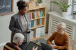 © Seventyfour - Young adult Black woman standing and holding digital tablet while talking to senior Caucasian man and senior Caucasian woman sitting at table, using laptops in classroom setting