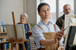 © Seventyfour - Middle aged Caucasian woman painting on canvas in classroom setting, standing in foreground with focused expression, senior Caucasian woman and senior Caucasian man painting in background