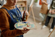 © Seventyfour - Middle aged Caucasian woman holding paint palette and brush demonstrating painting technique in classroom setting with blurred students in background participating in art lesson