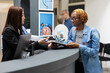 © DC Studio - Black woman registers for medical checkup as asian receptionist helps complete healthcare forms. African american patient listens as hospital administrator explains documentation at front desk.