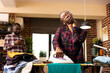 © DC Studio - Young woman looking tired and stressed while ironing shirt at home, standing at ironing board surrounded by laundry. African american husband lounges with smartphone, showing unequal household chores.
