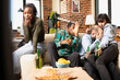 © DC Studio - Group of friends seated on couch, holding beer bottles and reacting with terrified expressions. Diverse individuals cover faces while black woman hides mouth, watching scary movie scene at home.