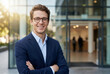 © ViskBx - Confident young businessman smiling with arms crossed outside modern office building