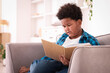 © New Africa - Boy reading book on sofa at home