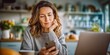 © NovaVision - Middle-aged woman looking at her smartphone while sitting at a table with a laptop