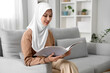 © Pixel-Shot - Beautiful young Muslim woman in hijab reading magazine and sitting on sofa in living room