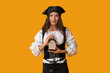© Pixel-Shot - Young woman in pirate costume holding bottle of rum isolated on orange background, halloween party character portrait in studio