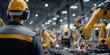 © NovaVision - Factory worker in yellow hard hat observes automated robotic arms welding on assembly line