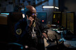 © pressmaster - Black young man wearing police uniform sitting at desk talking on phone using computer in dark office with monitors and documents visible in background