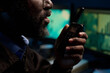 © pressmaster - Young Black man speaking into walkie talkie, holding device close to mouth, working in control room with computer monitors visible in background, focused on communication