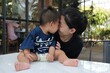 © anonpichit - Asian mother gently touching foreheads with her baby boy while sitting at an outdoor café.