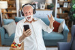 © Jelena Stanojkovic - Middle aged man joyfully dancing indoors wearing headphones holding a smartphone enjoying music and expressing happiness in a modern home.