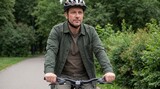 Middle-aged man wearing a bicycle helmet rides a mountain bike along a paved path surrounded by lush green trees and shrubs in a park setting