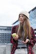© wavebreak3 - Woman wearing beanie and plaid shirt, leaning on concrete ledge in city holding apple and phone