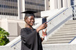 © wavebreak3 - African American woman wearing gown cap posing on campus holding phone selfie-stick and diploma