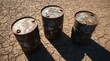 © Alex Pios - Three weathered metal oil barrels stand on dry, cracked earth under strong sunlight. Long shadows and peeling surfaces emphasize themes of pollution, resource extraction, and environmental impact