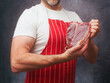 © mark_gusev - Athletic male butcher in white shirt and red and white classic stripe apron showing top quality T-bone steak in vacuum package for extra freshness of the product. Top price beef promotion.