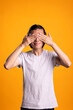 © DC Studio - Smiling man covering eyes with hands, pretending not to look at something. Playful male individual reacting with humor and shyness, expressing fun and cheerful attitude in yellow background studio.
