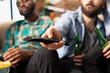 © DC Studio - White bearded man operating television remote, changing volume and channels, with friends sitting on couch close by. Male individual uses device in picking show to watch during casual hangout at home.