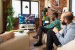 © DC Studio - Diverse young adults enjoying multiplayer console game at brick wall home. Friends relax on couch in front of television, watching and competing in fun gaming session in apartment.