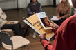 © pressmaster - Black woman holding psychotherapy support folder and informational leaflet while standing in group therapy session with multiethnic women sitting in background, focusing on mental health discussion